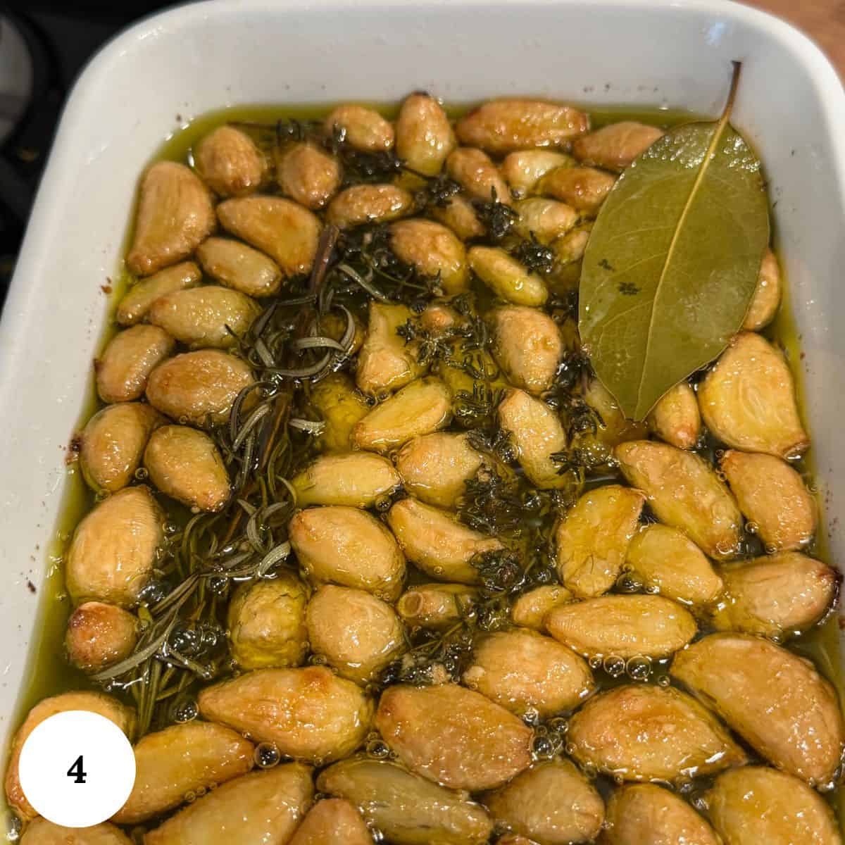 A white dish filled with garlic confit-roasted garlic cloves submerged in olive oil, garnished with fresh rosemary and a bay leaf.