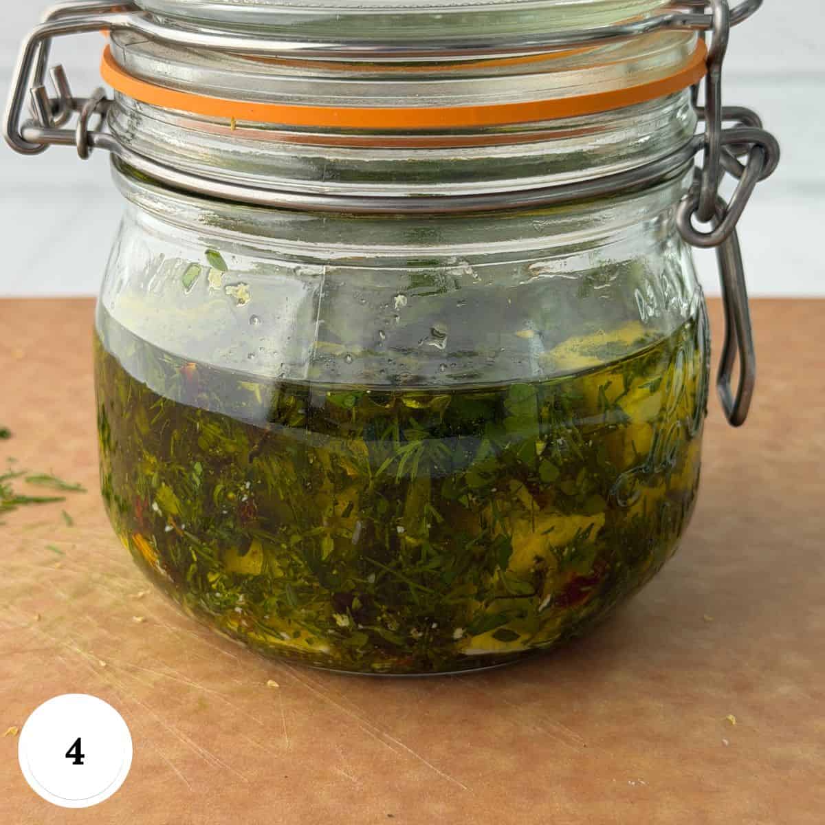 A glass jar with a metal clasp filled with chopped herbs, oil, and marinated feta cheese sits on a brown cutting board. The jar contains a green herb mixture in liquid. A white circle with the number 4 is in the corner.