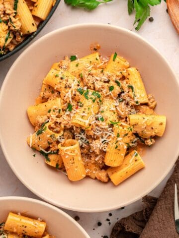 A bowl of rigatoni creamy pasta topped with ground meat, fresh herbs, and grated cheese, placed on a light table with bowls of pasta, grated cheese, and fresh parsley nearby.