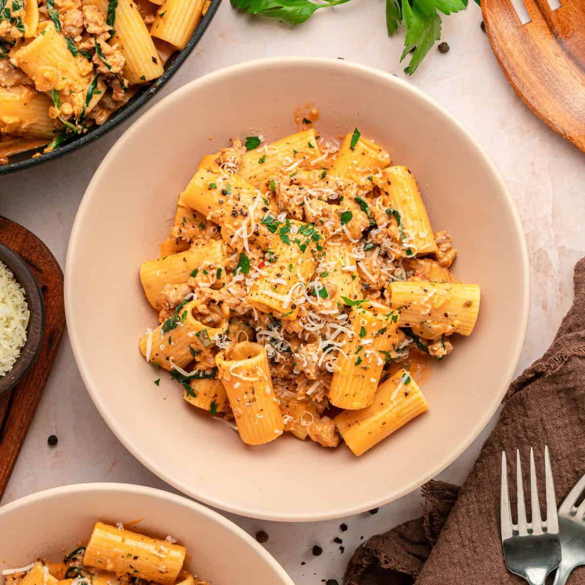 A bowl of rigatoni creamy pasta topped with ground meat, fresh herbs, and grated cheese, placed on a light table with bowls of pasta, grated cheese, and fresh parsley nearby.