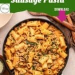 A skillet filled with creamy Italian sausage pasta garnished with herbs and parsley sits on a table beside grated cheese, garlic, black pepper, and spinach pasta. Text reads: Creamy Italian Sausage Pasta. Download.