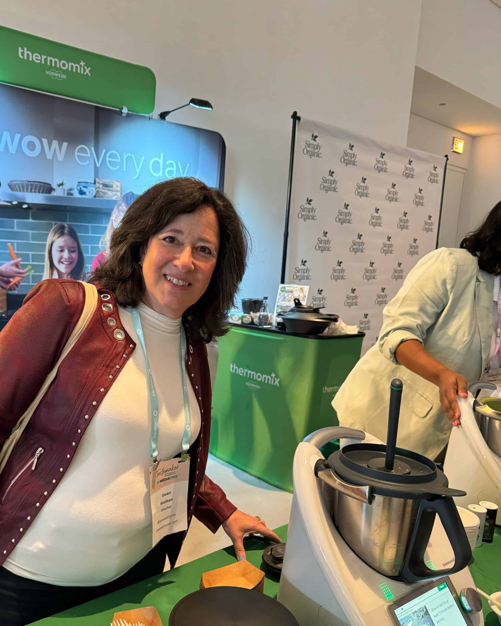 A woman in a red and white jacket smiles at a trade show booth displaying Thermomix kitchen appliances. Promotional banners and products are visible in the background.