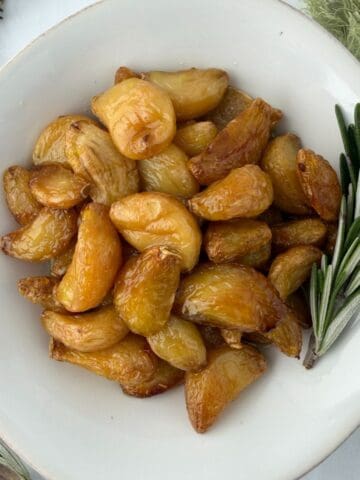 A white plate filled with golden brown garlic confit cloves, garnished with fresh rosemary sprigs on the side, placed on a light surface with a green cloth nearby.