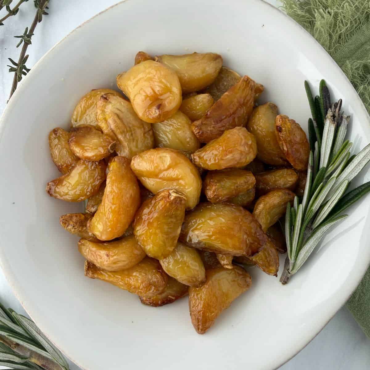 A white plate filled with golden brown garlic confit cloves, garnished with fresh rosemary sprigs on the side, placed on a light surface with a green cloth nearby.