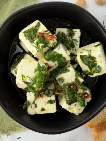 A black bowl filled with cubes of marinated feta cheese in herbs and oil, garnished with chopped parsley and red pepper. Slices of bread, a spoon, and a green napkin are nearby on a white surface.