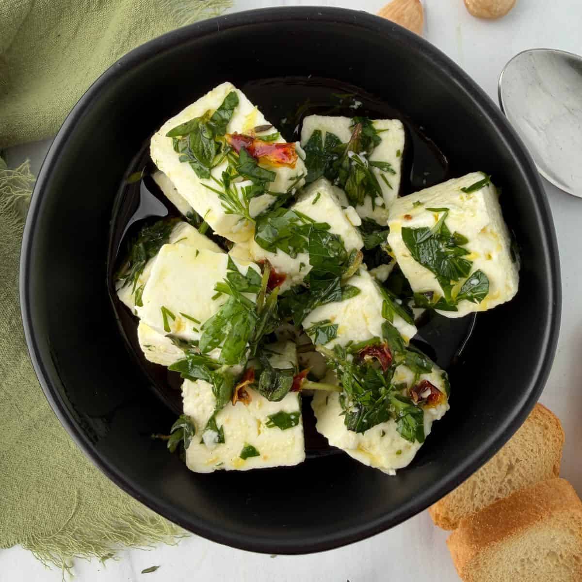 A black bowl filled with cubes of marinated feta cheese in herbs and oil, garnished with chopped parsley and red pepper. Slices of bread, a spoon, and a green napkin are nearby on a white surface.