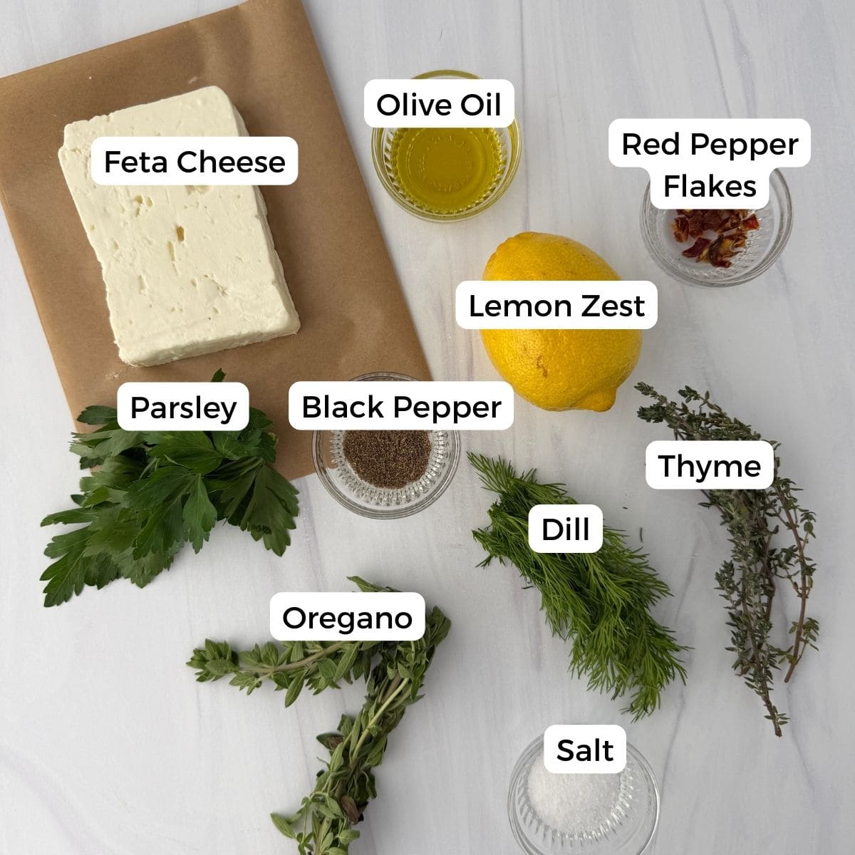 A flat lay of ingredients for marinated feta cheese on a white surface, including feta, parsley, oregano, dill, thyme, lemon, olive oil, red pepper flakes, black pepper, and salt—each labeled with text.