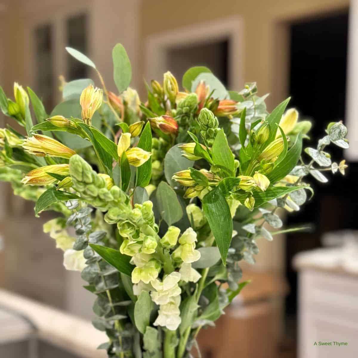 A close-up of a bouquet of green and yellow flowers with leafy stems from Sunday Thymes, set indoors against a softly blurred kitchen background.