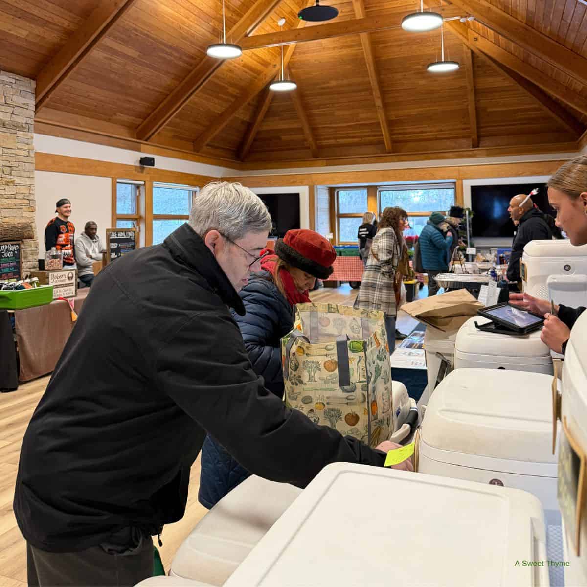 People browse and shop at the indoor Sunday Thymes farmers market with wooden ceilings. A man and woman examine goods at a vendor table while others mingle in the lively, warm atmosphere.