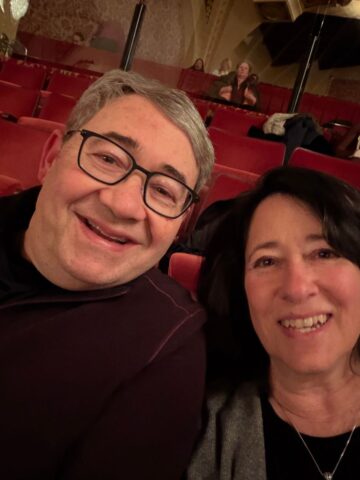 A man and woman smile for a selfie while sitting together in red theater seats at Sunday Thymes. Other people are seated in the background, holding drinks and chatting before a show.