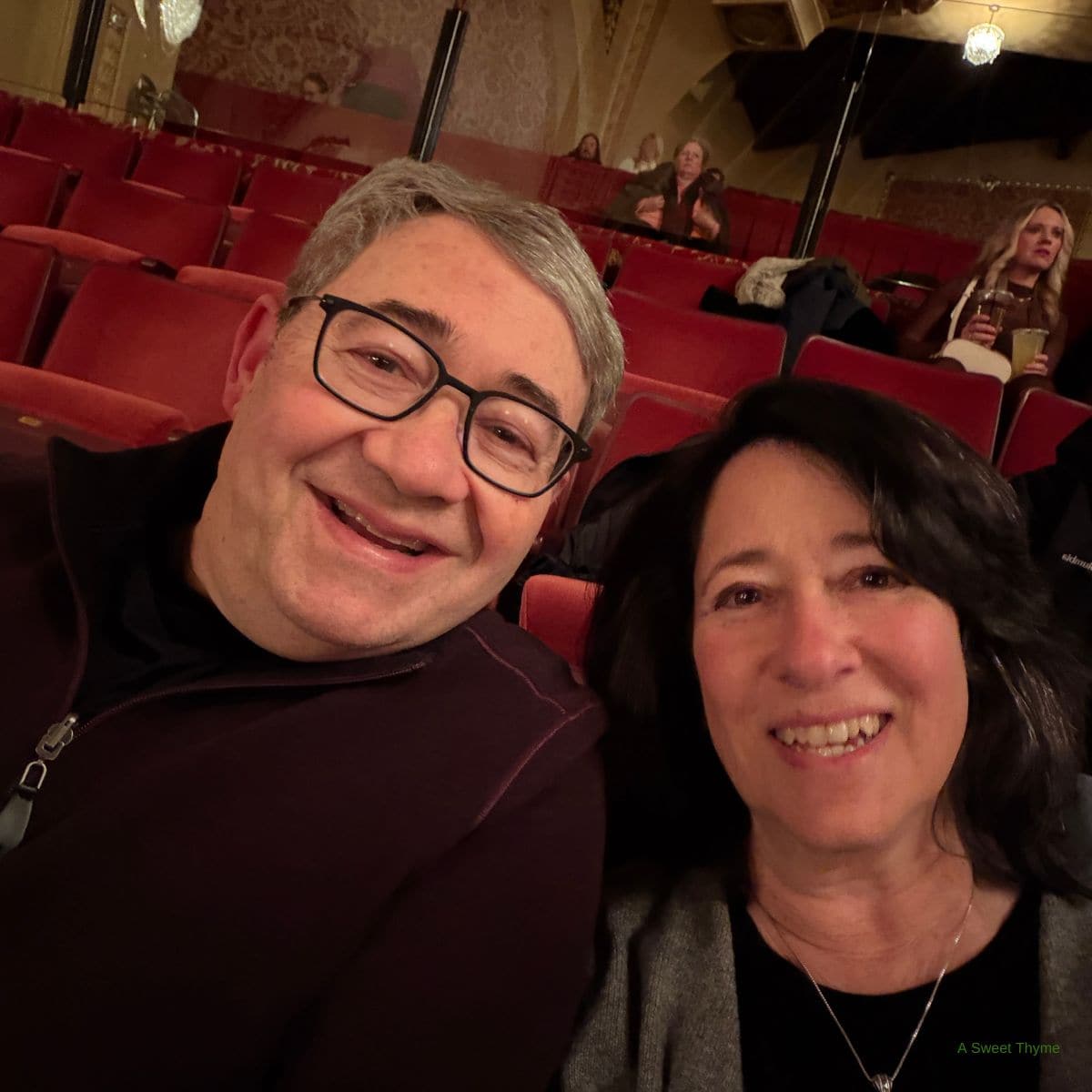 A man and woman smile for a selfie while sitting together in red theater seats at Sunday Thymes. Other people are seated in the background, holding drinks and chatting before a show.