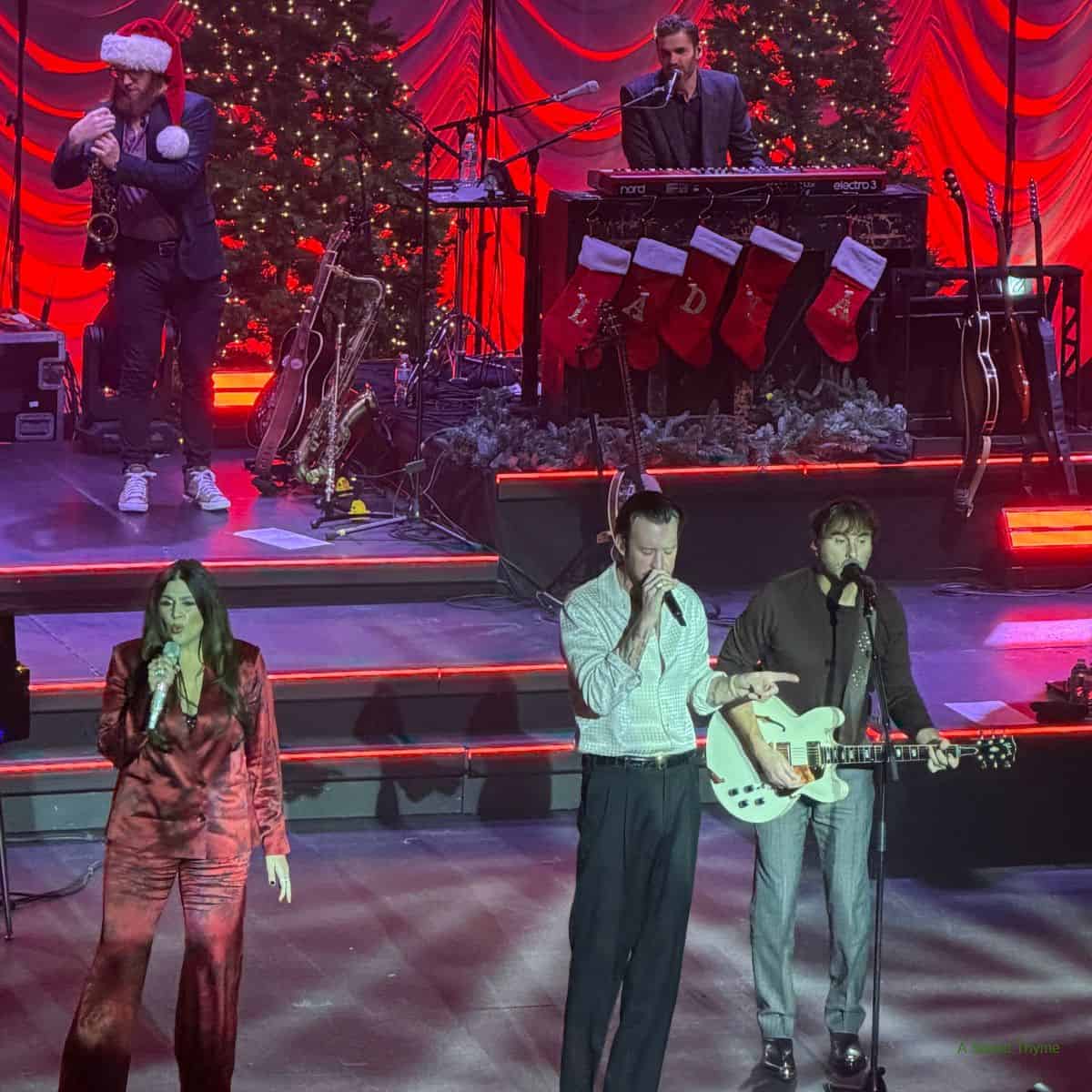 The Sunday Thymes perform onstage with festive Christmas decorations, including trees and stockings. Three singers stand at the front, one holding a guitar, while musicians play keyboards and saxophone in the background under red lighting.