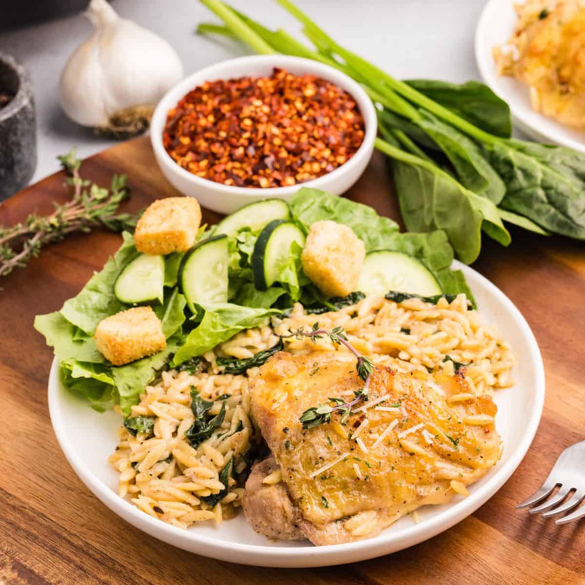 A plate of chicken orzo bake with crispy roasted chicken, served atop creamy orzo pasta with spinach, accompanied by a green salad with cucumber slices and croutons. Bowls of red pepper flakes and fresh herbs are nearby.