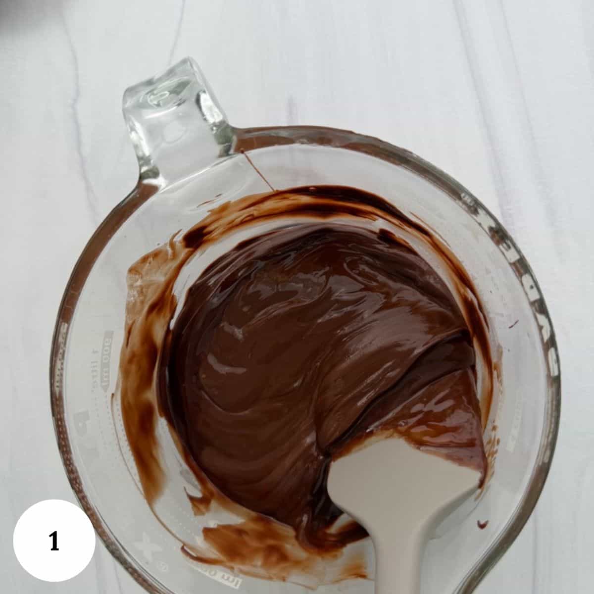 A glass measuring cup filled with melted chocolate, perfect for making chocolate krispy treats, is being stirred with a white spatula, seen from above. A white circle with the number 1 sits at the bottom left corner.