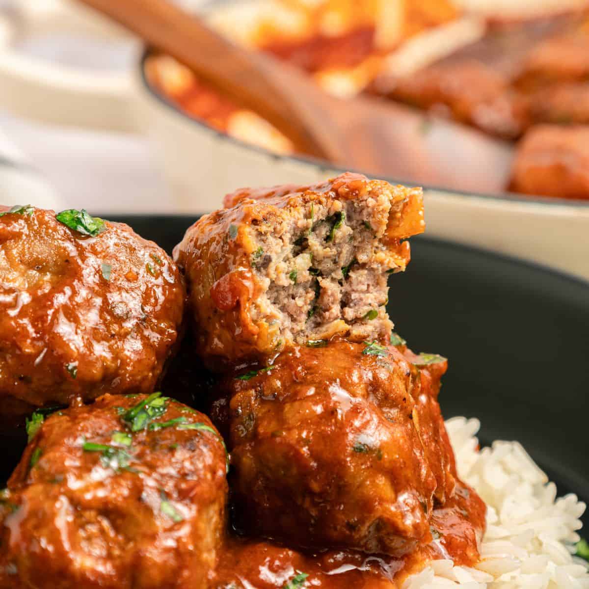 Three Greek beef meatballs, or Soutzoukakia, in a rich tomato sauce sit atop white rice in a black bowl. One meatball is cut open, revealing a moist, herbed interior. Fresh herbs are sprinkled on top, with more meatballs in the background.