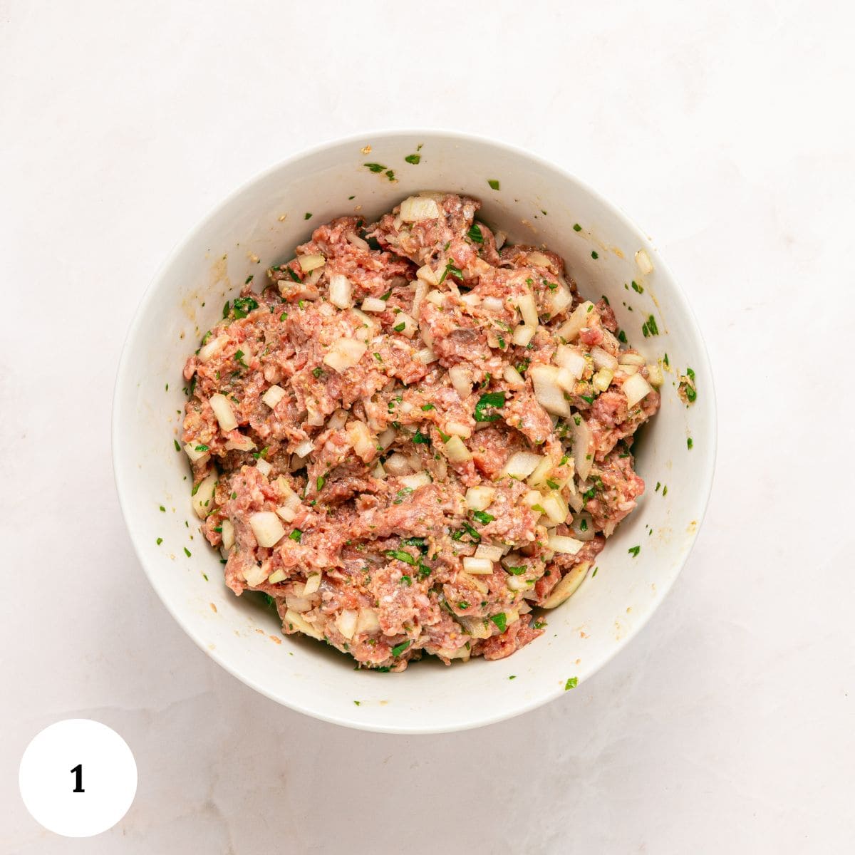 A white bowl filled with a mixture for Greek Beef Meatballs, combining ground meat, chopped onions, herbs, and seasonings, sits on a light-colored surface. A white circle with the number 1 is in the lower left corner.