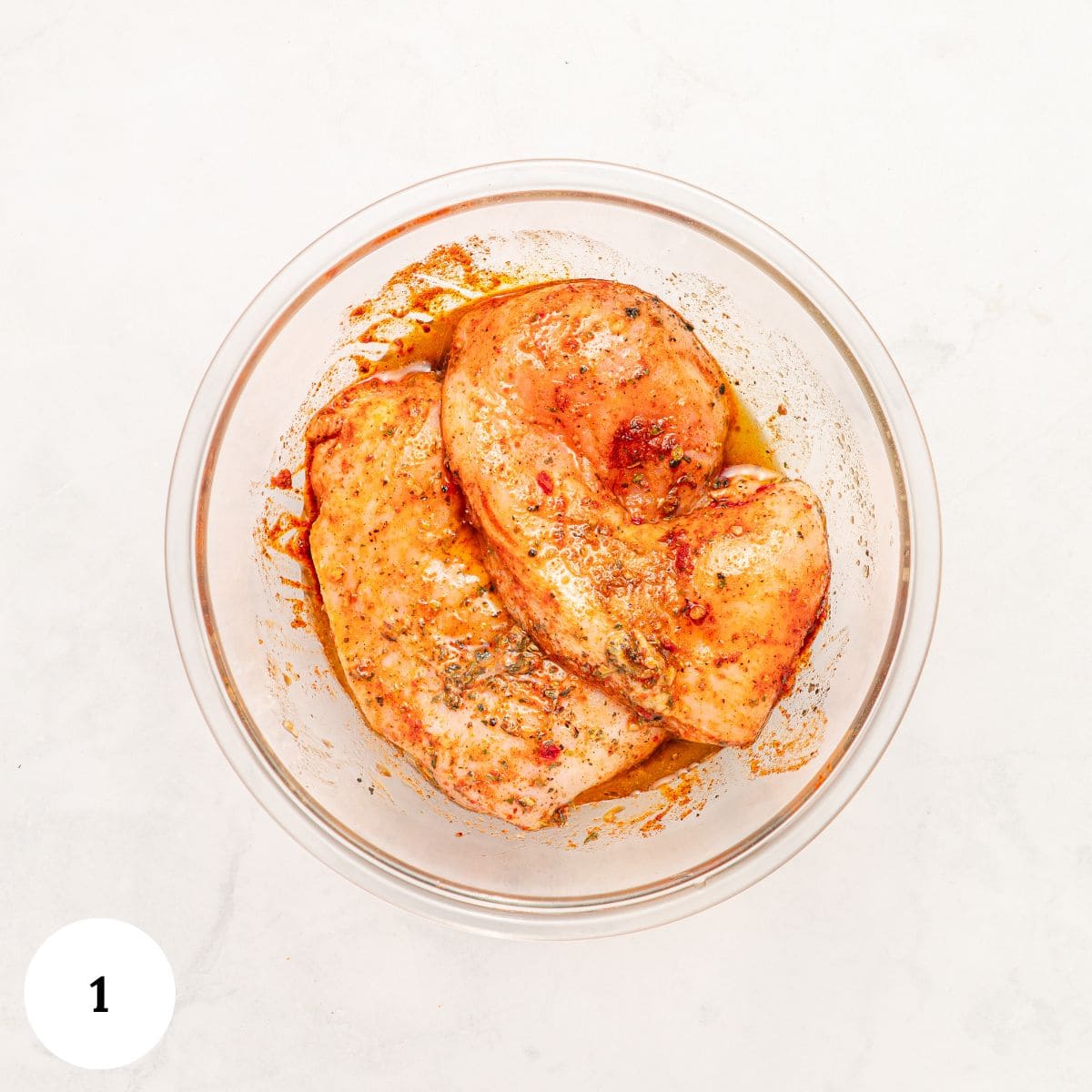 Raw, seasoned chicken breasts marinating in spices and oil inside a clear glass bowl, ready to become Mediterranean Chicken Couscous. The bowl sits on a white surface, with a white circle and the number 1 in the lower left corner.
