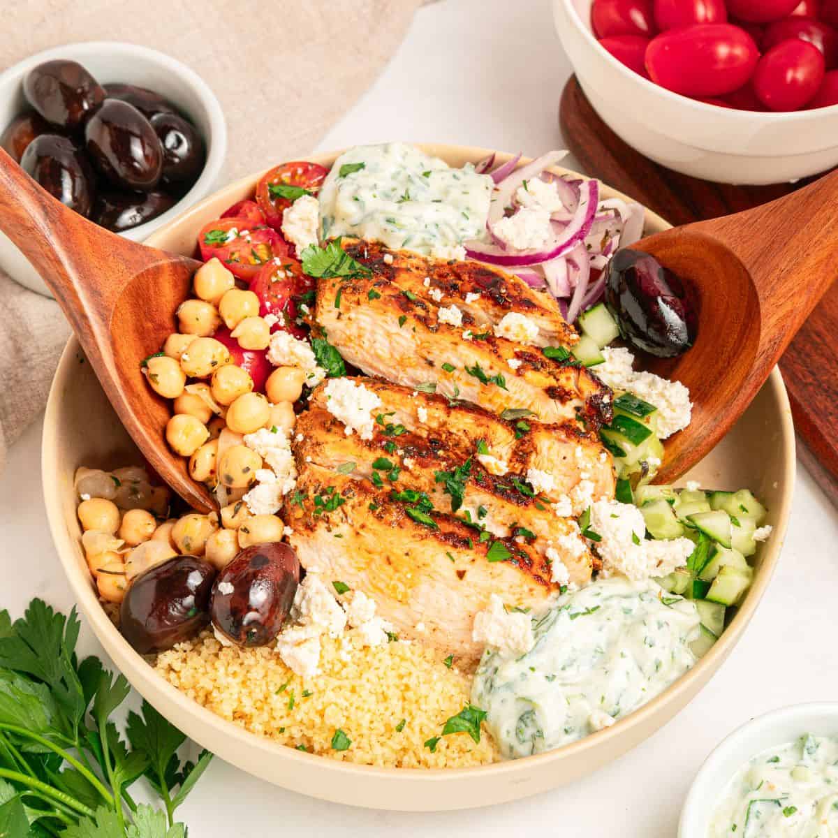 A vibrant Mediterranean chicken bowl featuring grilled chicken slices, chickpeas, kalamata olives, cherry tomatoes, cucumber, red onion, couscous, tzatziki sauce, and crumbled feta cheese, served with wooden salad servers.