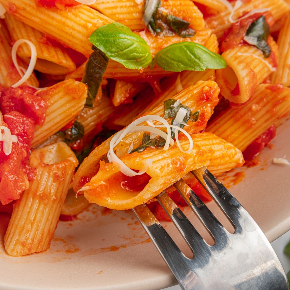A close-up of classic Italian Penne Pomodoro with rich Italian tomato sauce, grated cheese, and fresh basil leaves on a white plate. A fork holds a piece of pasta in the foreground.