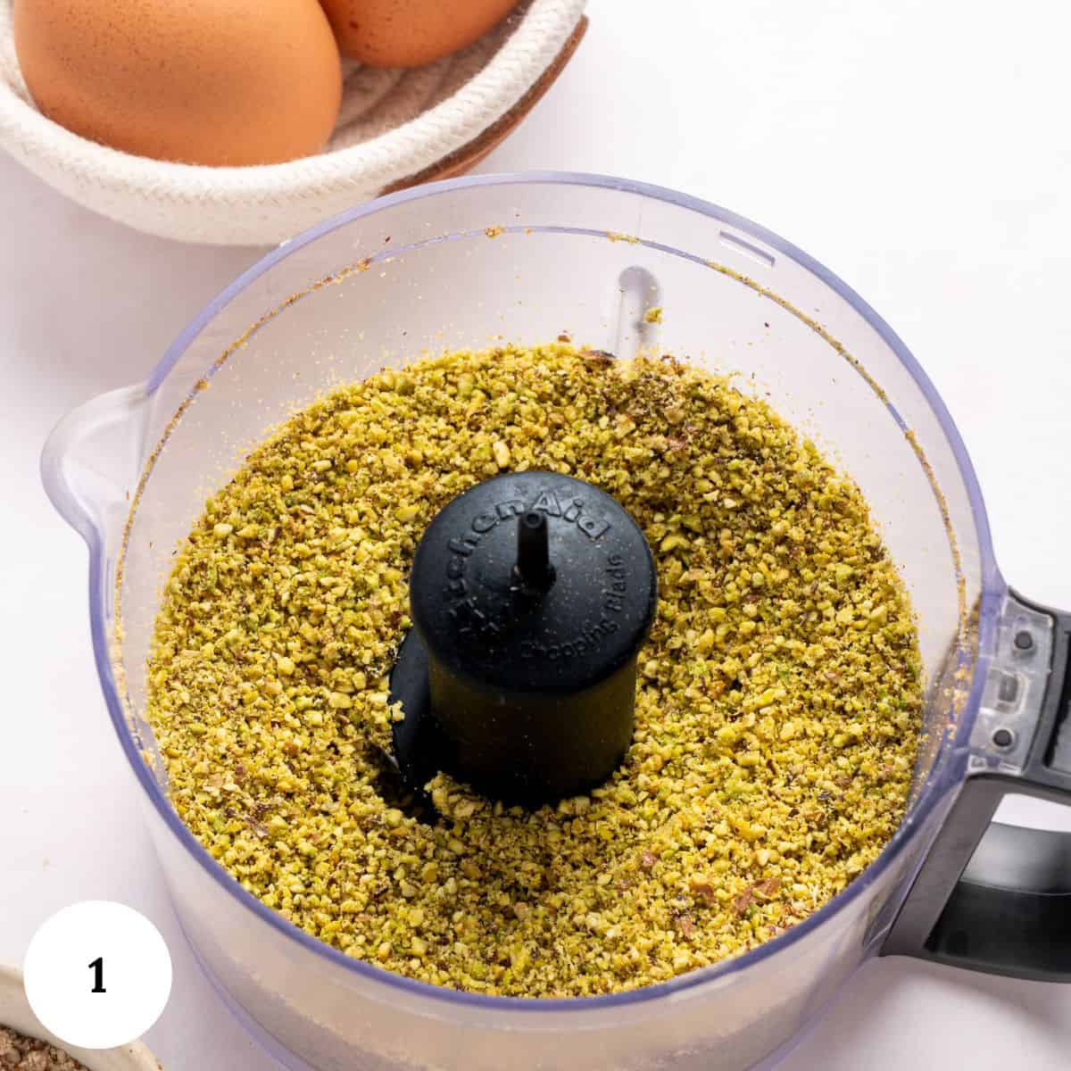 Crushed pistachios in a food processor bowl, with whole eggs in a small bowl nearby, ready to be used for making a pistachio ricotta cake, viewed from above on a white surface.