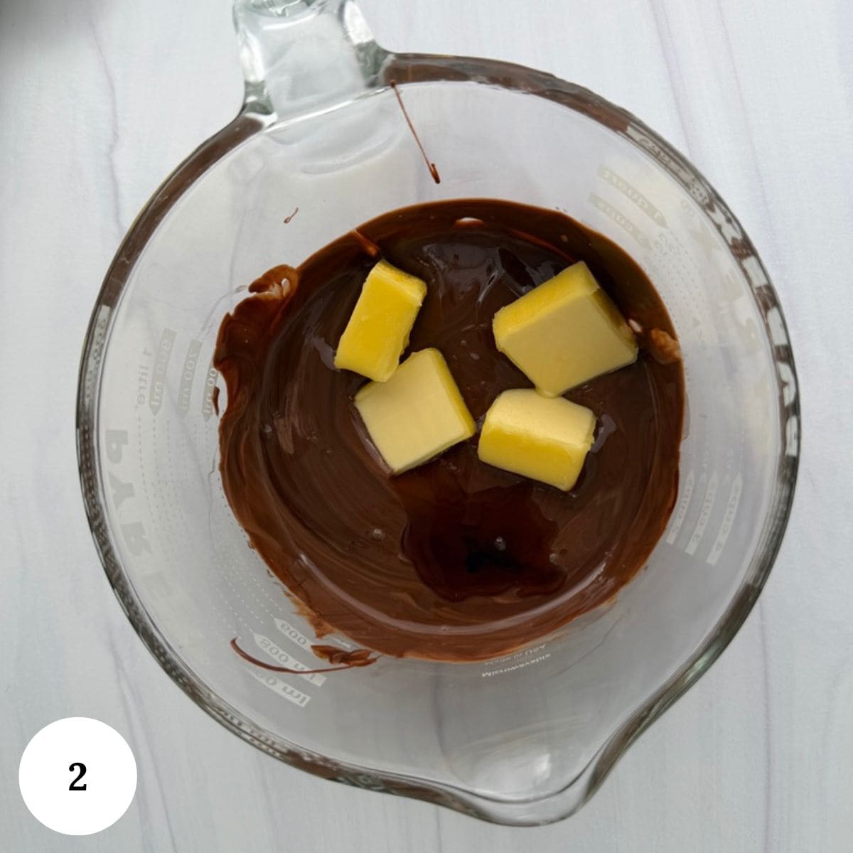 A glass measuring cup with melted chocolate and four cubes of butter inside, perfect for making chocolate krispy treats, sits on a white surface. A white circle with the number 2 is in the bottom left corner.
