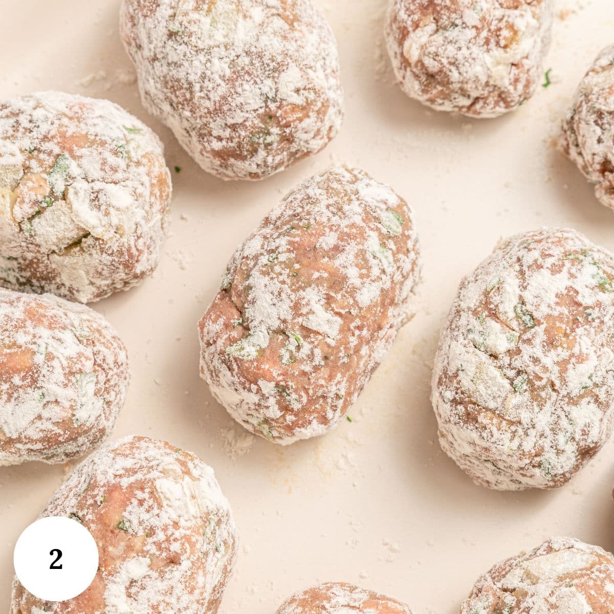 Several uncooked Greek beef meatballs coated in flour are arranged on a light-colored surface. There are visible green herb pieces in the soutzoukakia mixture. A small white circle with the number 2 is in the lower left corner.