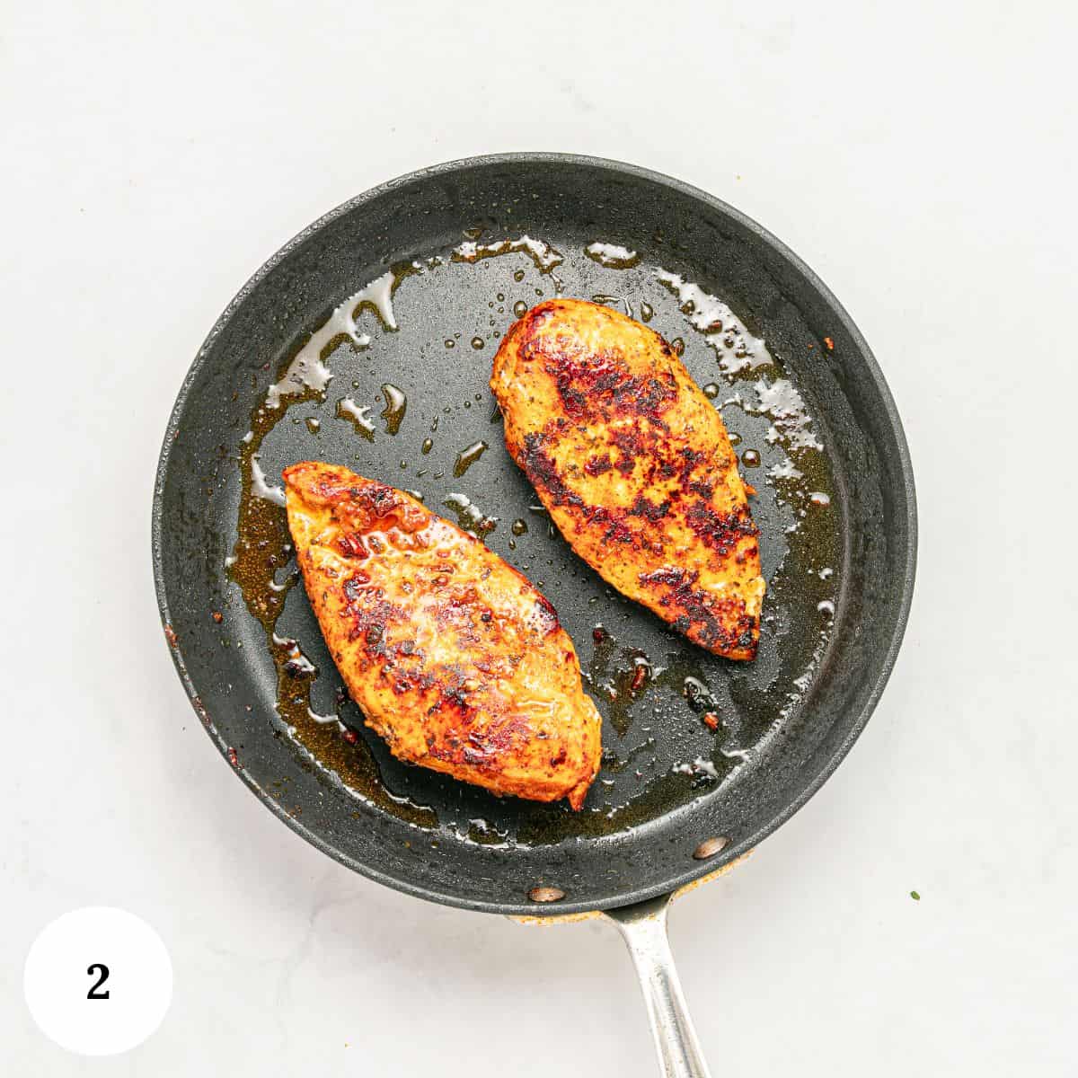 A nonstick frying pan with two cooked, browned chicken breasts on a white surface-perfect for a Mediterranean-inspired meal. A small circle with the number 2 is in the lower left corner.