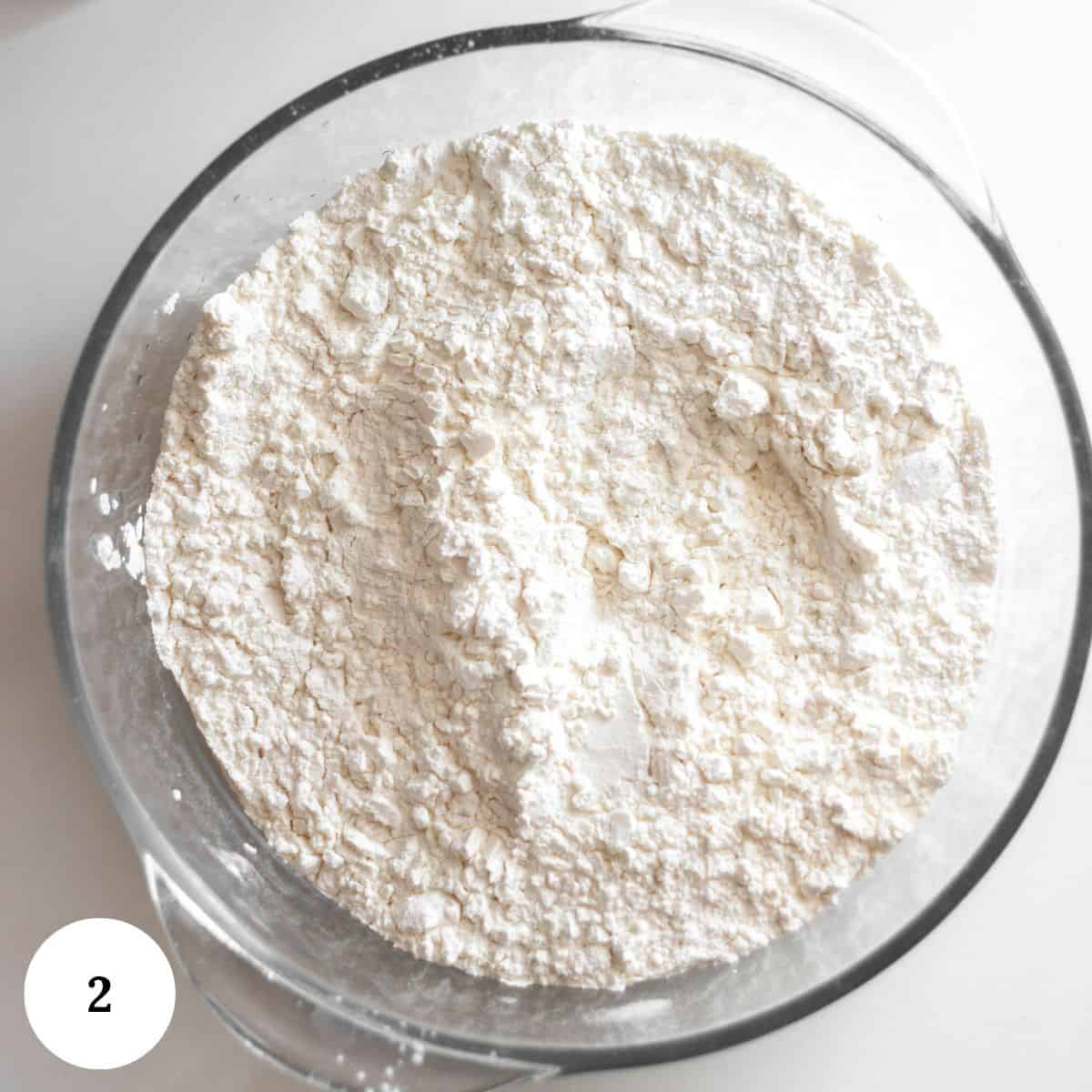 A clear glass bowl filled with white flour on a white surface, perfect for baking treats like pistachio ricotta cake. A small white circle in the bottom left corner displays the number 2.