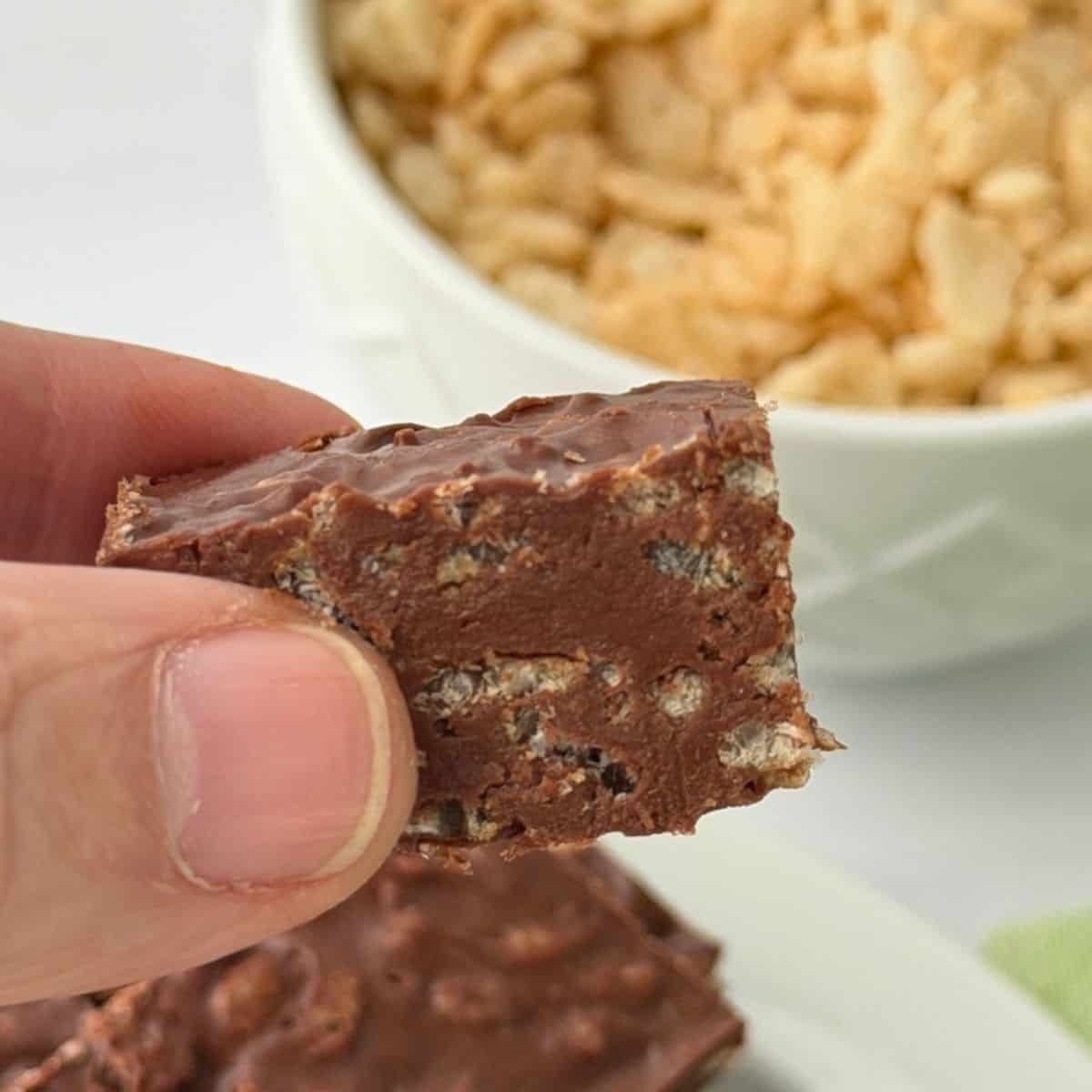 A hand holds a chocolate krispy treat with visible puffed rice pieces. In the background, a bowl filled with puffed rice is partially visible.