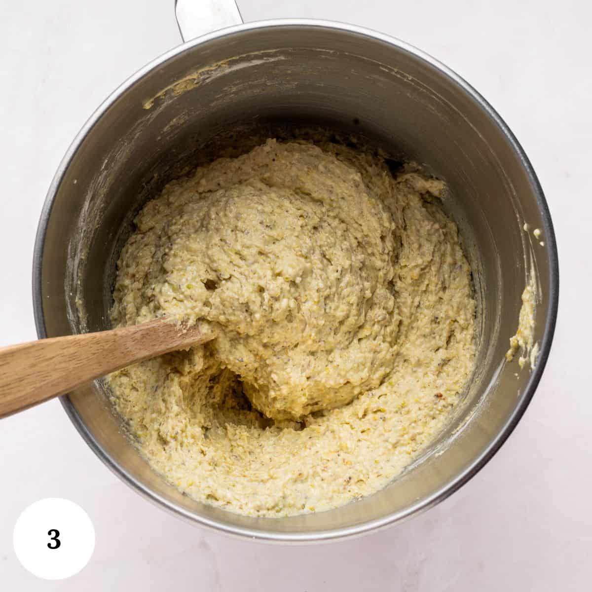 A mixing bowl filled with thick pistachio ricotta cake batter being stirred with a wooden spoon. The bowl is on a light surface, and a white circle with the number 3 is in the bottom left corner.
