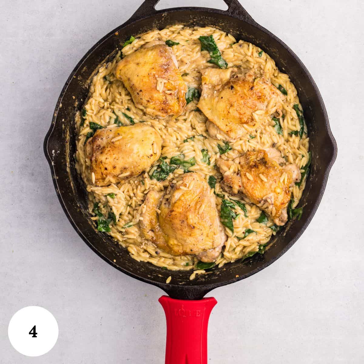 A skillet with creamy chicken orzo bake, wilted spinach, and four golden-brown chicken thighs, all cooked together. The skillet has a bright red handle and sits on a light gray surface, with a white circle marked 4 in the corner.