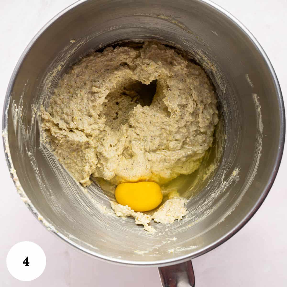A metal mixing bowl with dough and a raw egg on top, ready to be mixed for a pistachio ricotta cake. The bowl rests on a light surface, with a white circle and the number 4 in the bottom left corner.