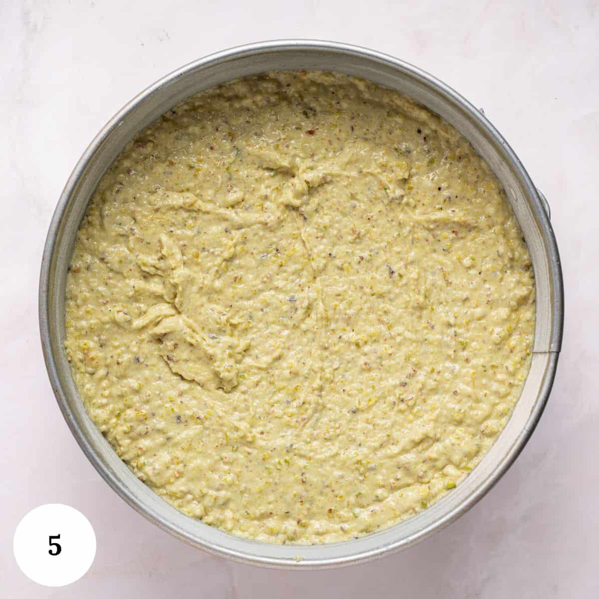 A round cake pan filled with unbaked, textured pistachio ricotta cake batter sits on a light-colored surface. A white circle with the number 5 appears in the lower left corner.