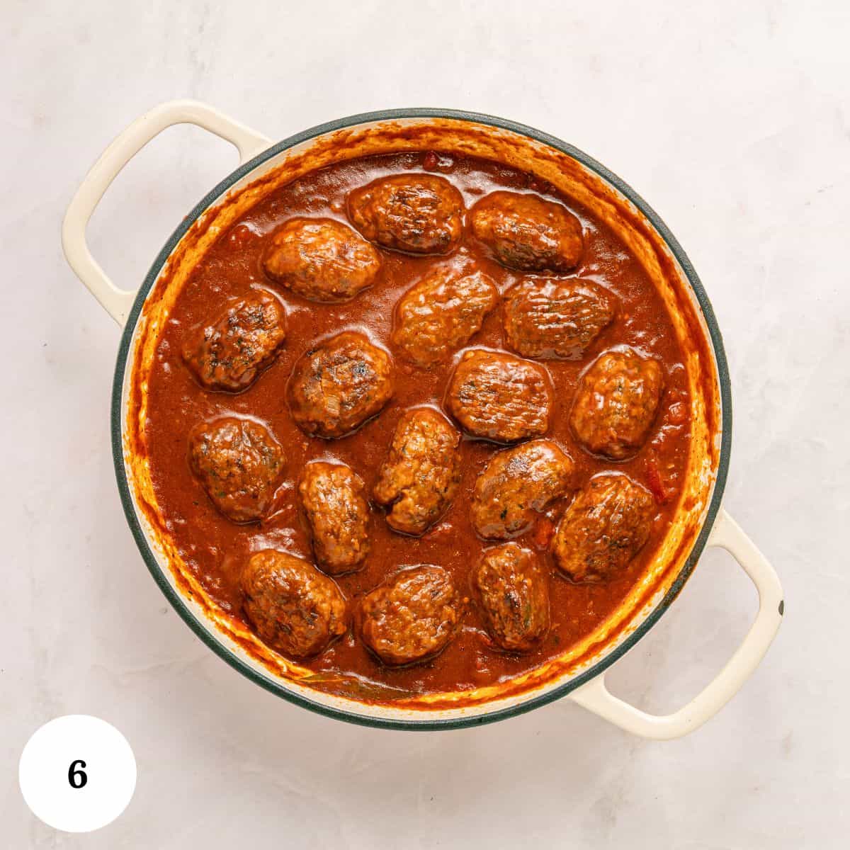 A round white pot filled with Greek Beef Meatballs, or Soutzoukakia, simmering in a rich red tomato sauce, viewed from above. There is a white circle with the number 6 in the lower left corner.