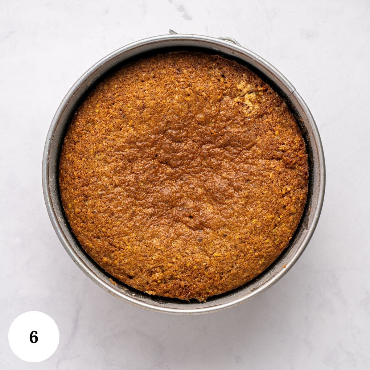A round, golden-brown pistachio ricotta cake in a metal pan sits on a light-colored surface. The number 6 appears in a white circle in the bottom left corner of the image.