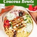 A vibrant couscous bowl featuring grilled chicken, chickpeas, cherry tomatoes, cucumbers, Kalamata olives, red onions, tzatziki sauce, and feta cheese—garnished with fresh herbs and a side of olives on a white surface.