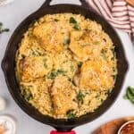 A cast iron skillet filled with creamy chicken orzo bake, wilted spinach, and five golden-brown chicken thighs, garnished with grated cheese and herbs, sits on a light surface near a striped towel.