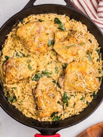 A cast iron skillet filled with creamy chicken orzo bake, wilted spinach, and five golden-brown chicken thighs, garnished with grated cheese and herbs, sits on a light surface near a striped towel.