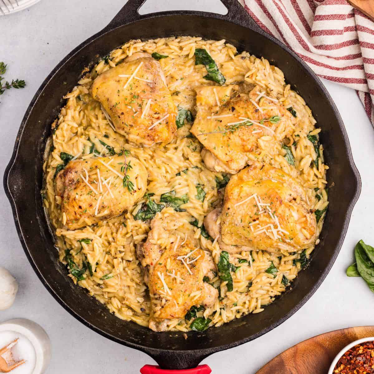 A cast iron skillet filled with creamy chicken orzo bake, wilted spinach, and five golden-brown chicken thighs, garnished with grated cheese and herbs, sits on a light surface near a striped towel.