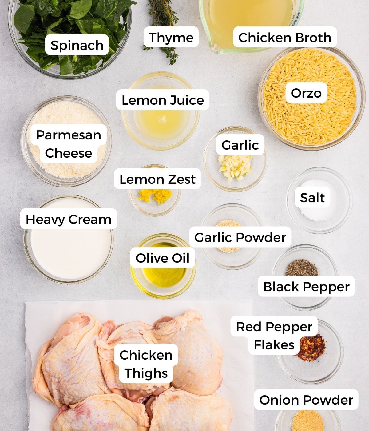 Ingredients for a chicken orzo bake arranged on a countertop, including spinach, thyme, chicken broth, orzo, Parmesan cheese, lemon juice and zest, garlic, salt, heavy cream, olive oil, spices, chicken thighs, and red pepper flakes.