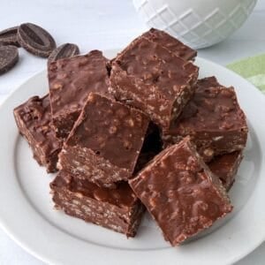 A white plate stacked with chocolate krispy treats, their crunchy, textured surface on display, sits on a white table near a few chocolate pieces and a partially visible white bowl.
