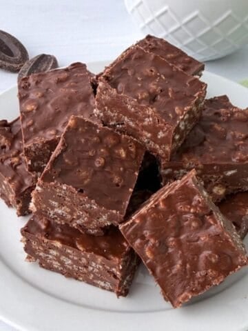 A white plate stacked with chocolate krispy treats, their crunchy, textured surface on display, sits on a white table near a few chocolate pieces and a partially visible white bowl.