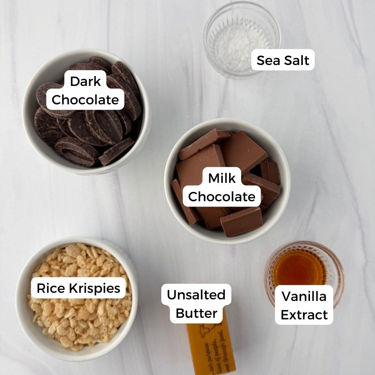 Five small bowls and a stick of butter are arranged on a white surface, each labeled: dark chocolate, milk chocolate, sea salt, rice krispies, unsalted butter, and vanilla extract—perfect for making delicious chocolate krispy treats.