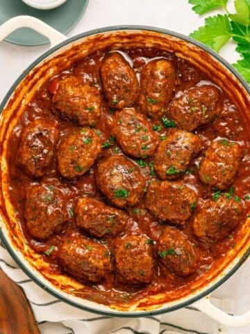 Round white pot filled with Greek Beef Meatballs, or Soutzoukakia, in rich red tomato sauce, garnished with chopped parsley. A striped towel, wooden spoon, and fresh parsley leaves are nearby on a light-colored tabletop.