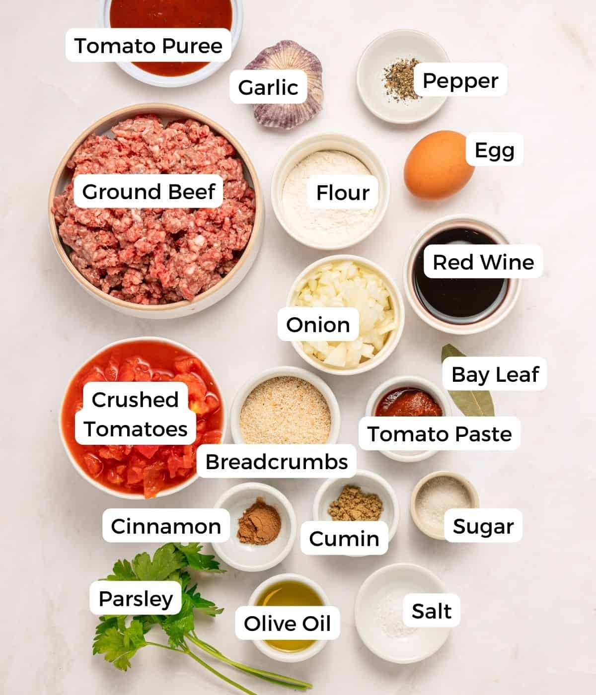 Top view of labeled ingredients for Soutzoukakia, including ground beef, tomato sauce, red wine, garlic, onion, egg, flour, breadcrumbs, tomato paste, parsley, olive oil, spices and seasonings—perfect for Greek beef meatballs.