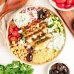 A Mediterranean bowl filled with grilled chicken, couscous, cherry tomatoes, chickpeas, cucumber, kalamata olives, red onion, tzatziki sauce, and crumbled feta cheese, surrounded by bowls of olives and tomatoes.