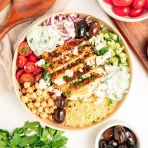 A Mediterranean bowl filled with grilled chicken, couscous, cherry tomatoes, chickpeas, cucumber, kalamata olives, red onion, tzatziki sauce, and crumbled feta cheese, surrounded by bowls of olives and tomatoes.