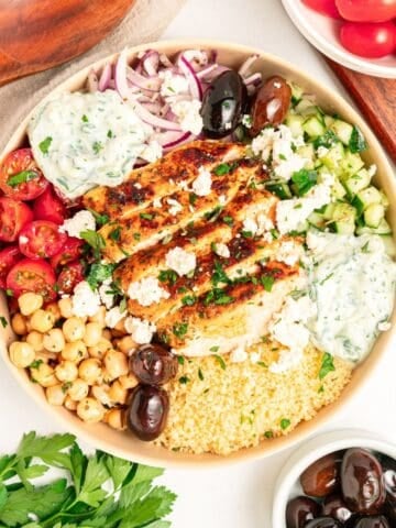 A Mediterranean bowl filled with grilled chicken, couscous, cherry tomatoes, chickpeas, cucumber, kalamata olives, red onion, tzatziki sauce, and crumbled feta cheese, surrounded by bowls of olives and tomatoes.