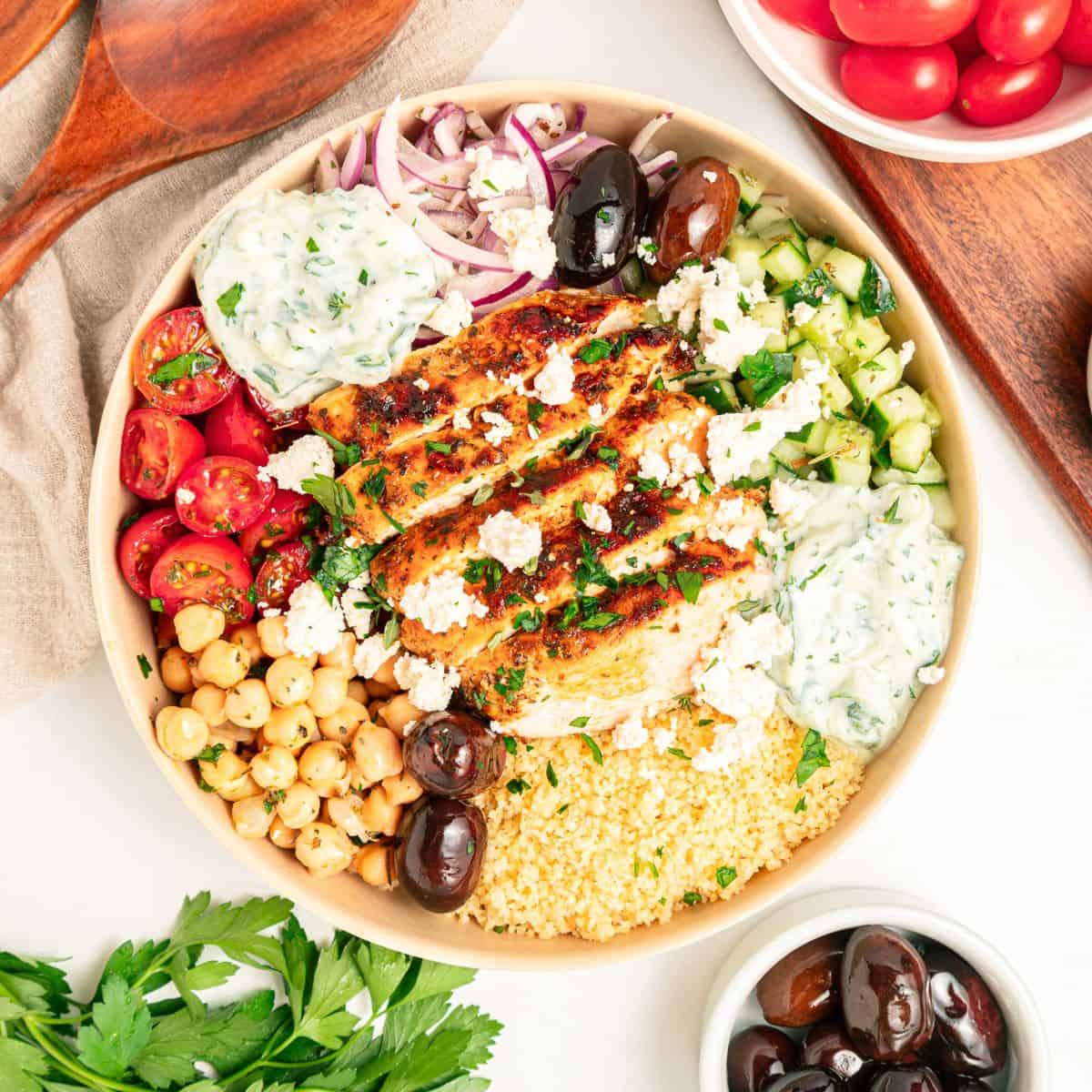A Mediterranean bowl filled with grilled chicken, couscous, cherry tomatoes, chickpeas, cucumber, kalamata olives, red onion, tzatziki sauce, and crumbled feta cheese, surrounded by bowls of olives and tomatoes.