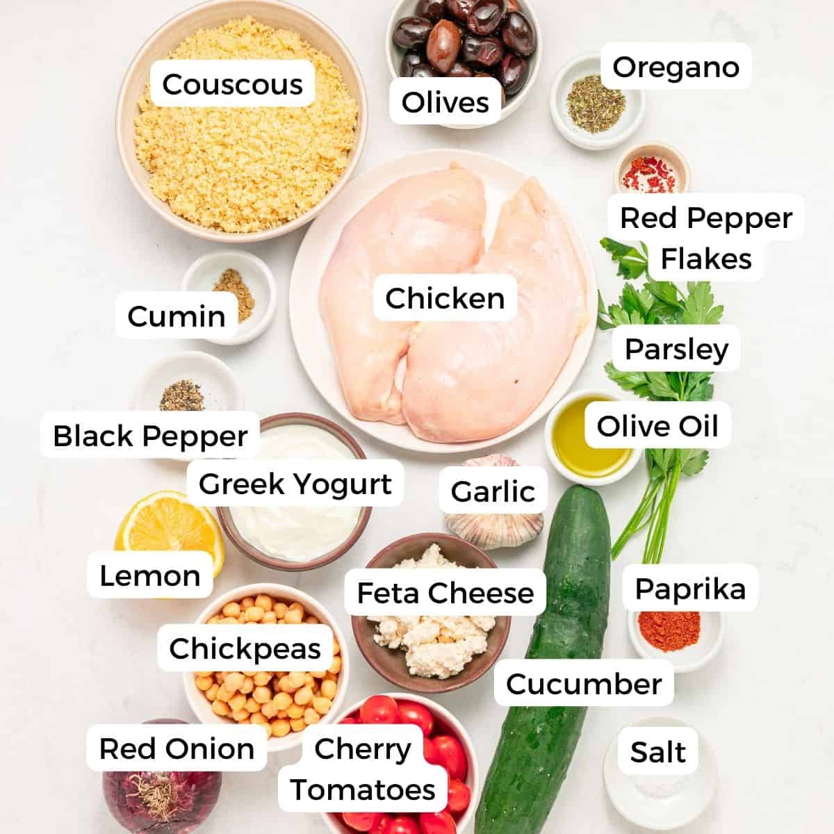 A flat lay of labeled ingredients for Mediterranean couscous bowls, including chicken, olives, Greek yogurt, chickpeas, feta cheese, cucumber, cherry tomatoes, red onion, garlic, lemon, parsley, olive oil, and spices.